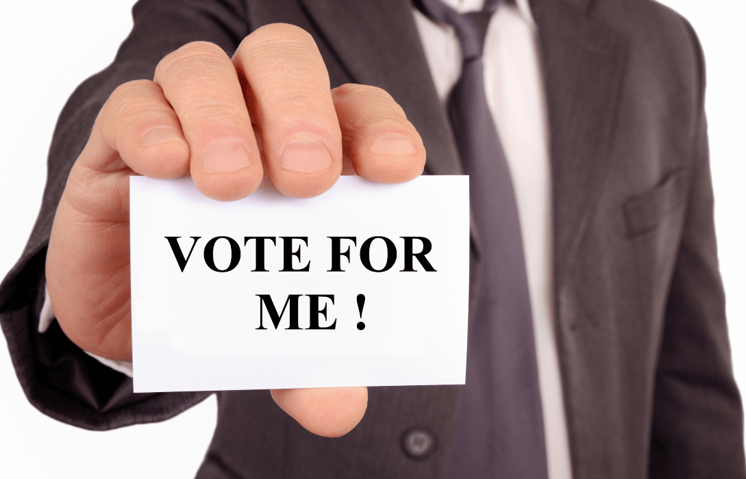 Close-up of a businessman in a suit holding a white card that says VOTE FOR ME in bold black letters for Cumhuritey.