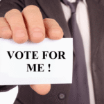 Close-up of a businessman in a suit holding a white card that says VOTE FOR ME in bold black letters for Cumhuritey.