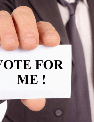 Close-up of a businessman in a suit holding a white card that says VOTE FOR ME in bold black letters for Cumhuritey.