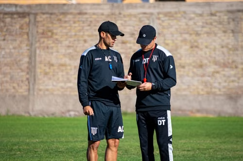Two football coaches reviewing training notes while wearing the Forest Command Coach Polo on the practice field.